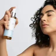 Person holding a bottle of herbal toner, eyes closed, curly hair.