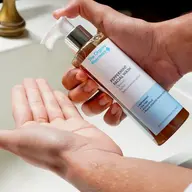 Person dispensing peppermint facial wash into hand by a sink.