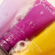 Three Caudalie shower gels in purple, pink, and yellow, covered in bubbles.