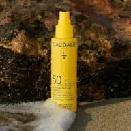 Yellow Caudalie SPF 50 spray bottle on a sandy beach with rocks in the background.