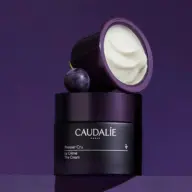 Caudalie Premier Cru cream jar with lid open, next to a grape, on a purple background.