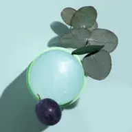 Green jar with blue cream, eucalyptus leaves, and a purple plum on a light blue background.