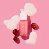 Pink Caudalie serum bottle with grapes and cream blobs on pink background.
