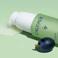 Caudalie Vinopure bottle with pump, liquid spill, and a grape on a green background.