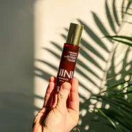 Hand holding NINI Organics Crimson Beauty serum, with plant shadow in background.