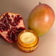 Open jar of cream with pomegranate half and whole mango on beige background.