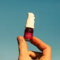 Hand holding a small skincare bottle with a pump against a clear blue sky.