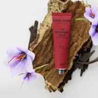 Red deodorant balm tube on bark with purple flowers. Text: Santal Nomad, AKT, saffron, sandalwood, leather.