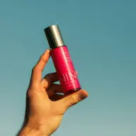 Hand holding pink skincare bottle against clear blue sky.