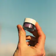 Hand holding a small jar of purple cream against a clear blue sky.