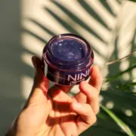 Hand holding a jar of NINI Organics violet skincare gel, with palm leaf shadows.