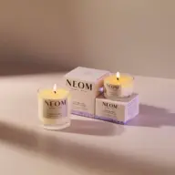 Two lit NEOM candles on a table with branded boxes in soft lighting.