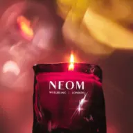 Red candle with flame, NEOM Wellbeing London text, blurred colourful background.