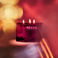 Red NEOM candle with three flames, blurred warm background. Text: "Wellbeing | London".