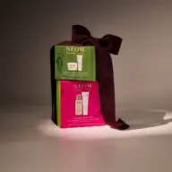 Gift box with NEOM skincare products, green and pink packaging, tied with a dark purple ribbon.