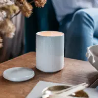 Lit white candle in a ceramic holder on a wooden table, with a lid beside it.