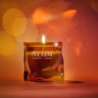 Lit NEOM candle with amber glass, text "Wellbeing | London", warm bokeh background.
