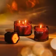Three lit NEOM candles in amber glass jars on a warm-toned background.
