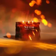 Lit candle in a dark amber glass with "NEOM Wellbeing London" text, warm glowing bokeh background.