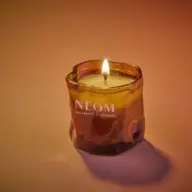 Lit candle in amber glass holder with "NEOM Wellbeing London" text. Warm background.