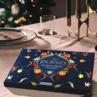 Tea advent calendar on a dining table with lit candles and a Christmas tree in the background.