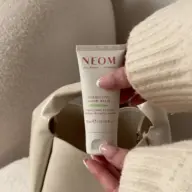 NEOM Energising Hand Balm, 30ml, held by a person in a cream jumper, near a beige bag.