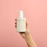 Hand holding NEOM hand lotion bottle against pink background.