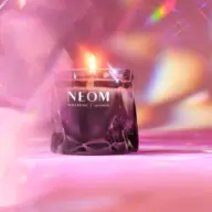 Lit candle in a glass holder with "NEOM Wellbeing London" text, pink and colourful background.