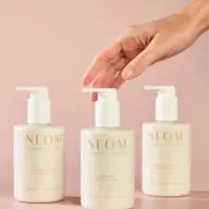 Three NEOM hand lotions on pink background, hand reaching for centre bottle.