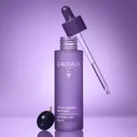 Purple Caudalie Anti-Hair Loss Serum bottle with dropper and grape on purple background.