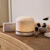 White NEOM diffuser on a wooden table with essential oils and packaging.