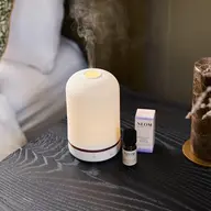 White diffuser emitting mist, with NEOM essential oil and box on a dark wooden table.