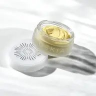 Open jar of yellow cream with "OSKIA" on the side, lid beside it on a white surface.