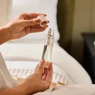 Hands holding a NEOM sleep spray bottle with cap removed, near a bed.