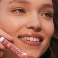 Smiling person holding RMS Beauty lip gloss tube. Text: "RUMOR".