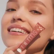 Smiling person holding a tube of RMS Beauty lip gloss labelled "Crush".