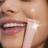 Close-up of a smiling person holding a pink tube labelled "rms" near their face.