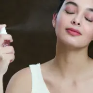 Woman spraying facial mist with eyes closed, wearing a white top.