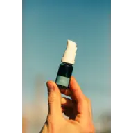 Hand holding a small blue-labelled spray bottle against a clear sky.