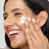 Woman applying cream to her cheek, smiling, with neatly manicured nails.