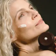 Woman applying cream to cheek, holding brown jar.