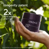 Hand holding Caudalie Premier Cru cream jar, green leaves in background. Text: "Longevity patent, 2x more volume after 1 j...