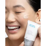 Smiling person applying The Organic Pharmacy Enzyme Peel Mask to cheek.