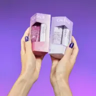 Two hands holding Caudalie skincare gift sets against a purple background.