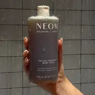 Hand holding Neom Feeling Grounded Body Wash, 500ml, in a tiled shower.