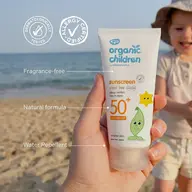Hand holding Organic Children SPF 50+ sunscreen on a beach. Child in background.