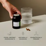 Hand holding Wild Nutrition Vitamin C Plus bottle, glass of water, two capsules on table.