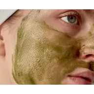 Close-up of a person's face with green clay mask applied, wearing a white headband.