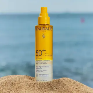 Caudalie SPF 50+ sun spray on sand with sea background.