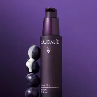 Caudalie Premier Cru Serum bottle with grapes and cream on purple background.